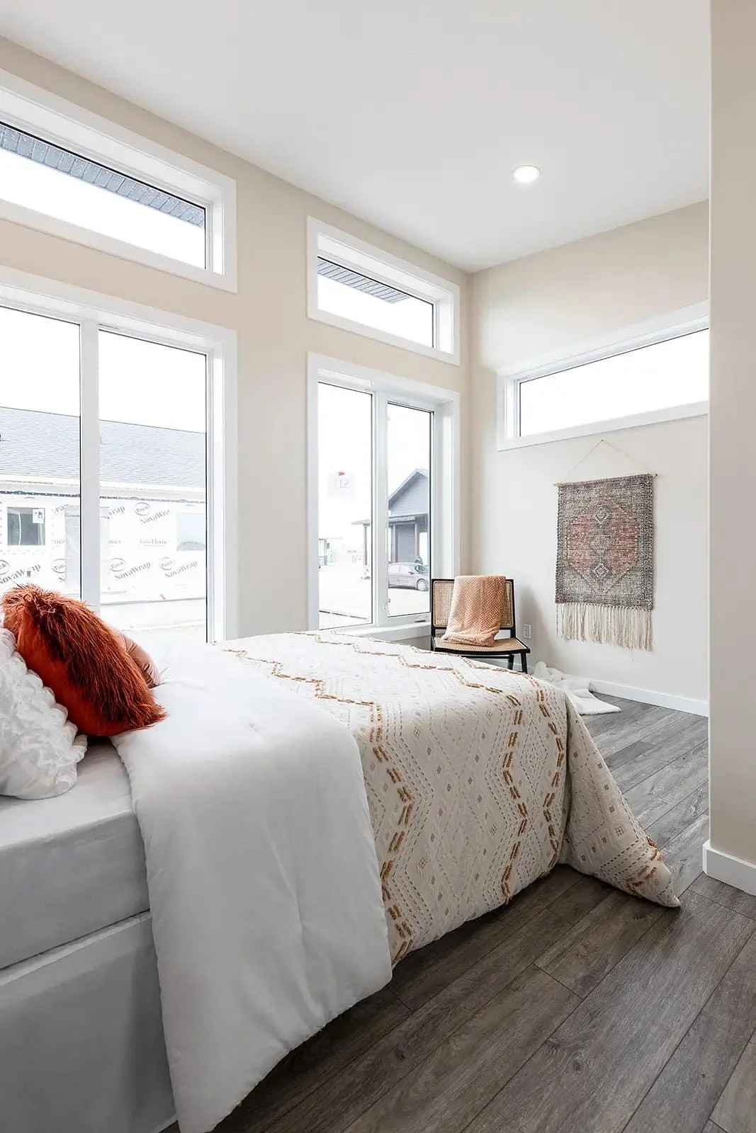 Bright bedroom with large windows, white walls, and wood flooring. Bed with textured blanket and orange cushions, chair with throw, and a woven wall hanging.