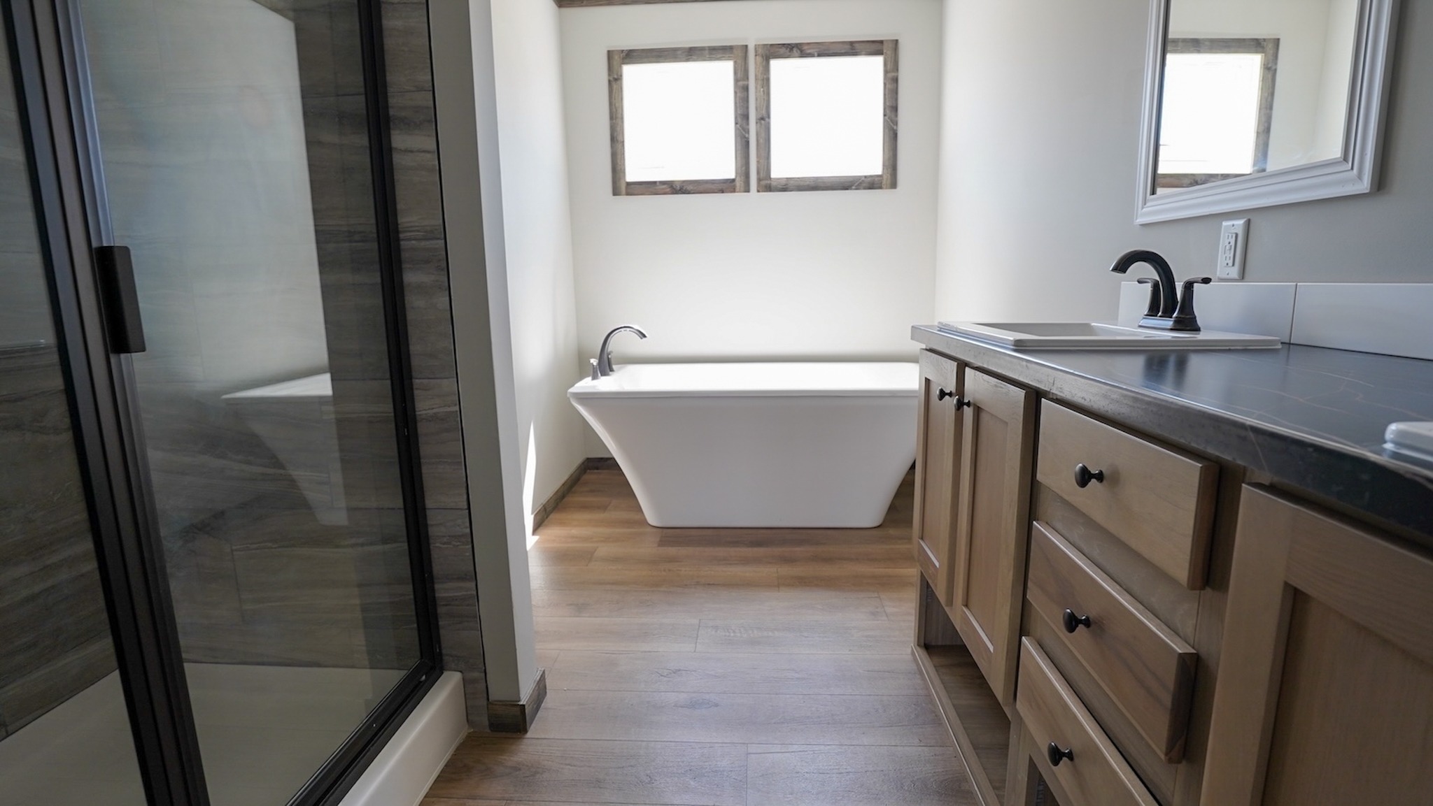 Modern bathroom with wooden floors, a sleek white bathtub beneath three small windows, a glass shower on the left, and a wooden vanity with a mirror.