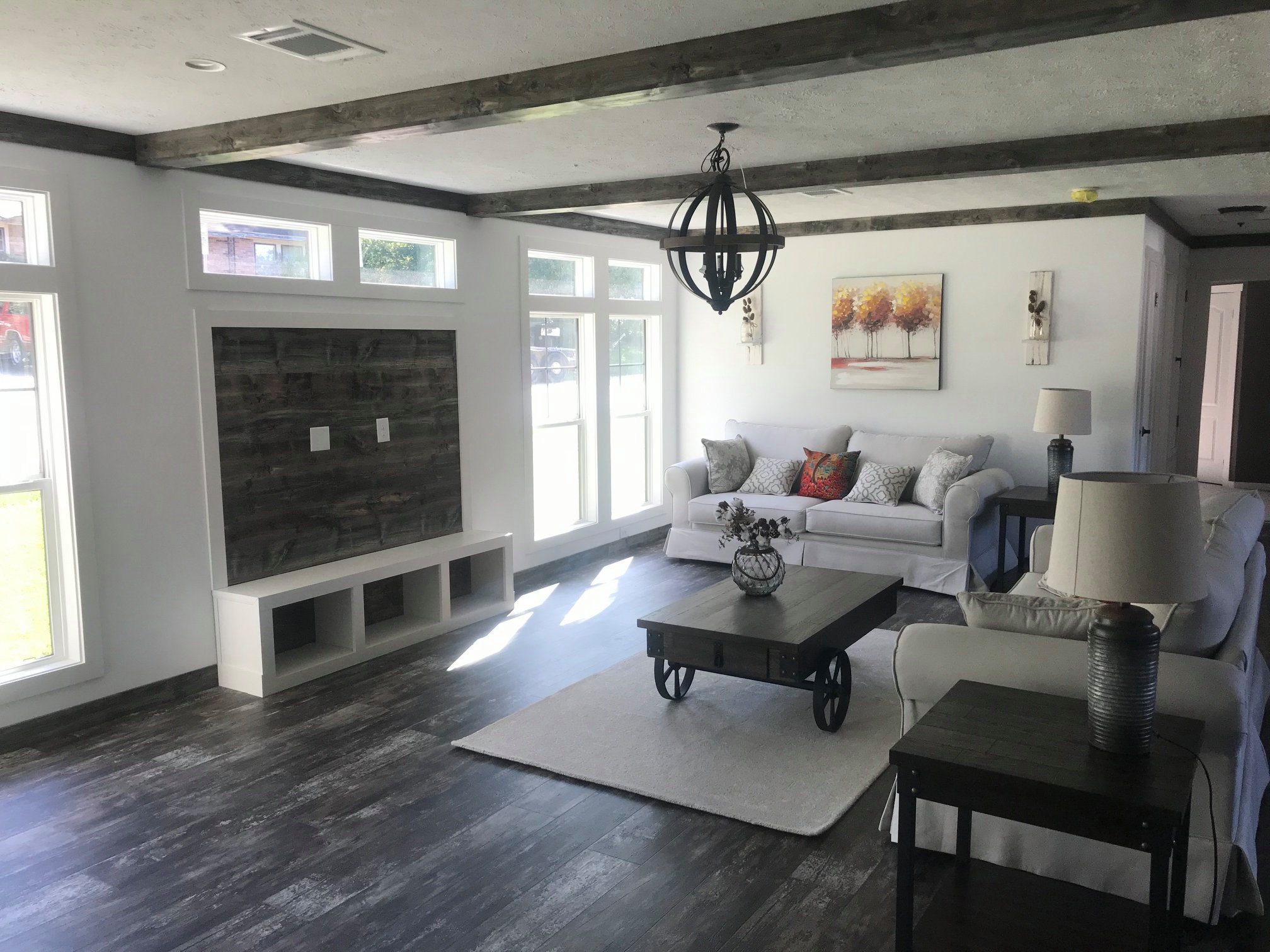 Spacious living room with white walls and dark wood floors. Features white sofas, rustic coffee table, modern chandelier, large windows, and landscape art. Bright, cozy ambiance.