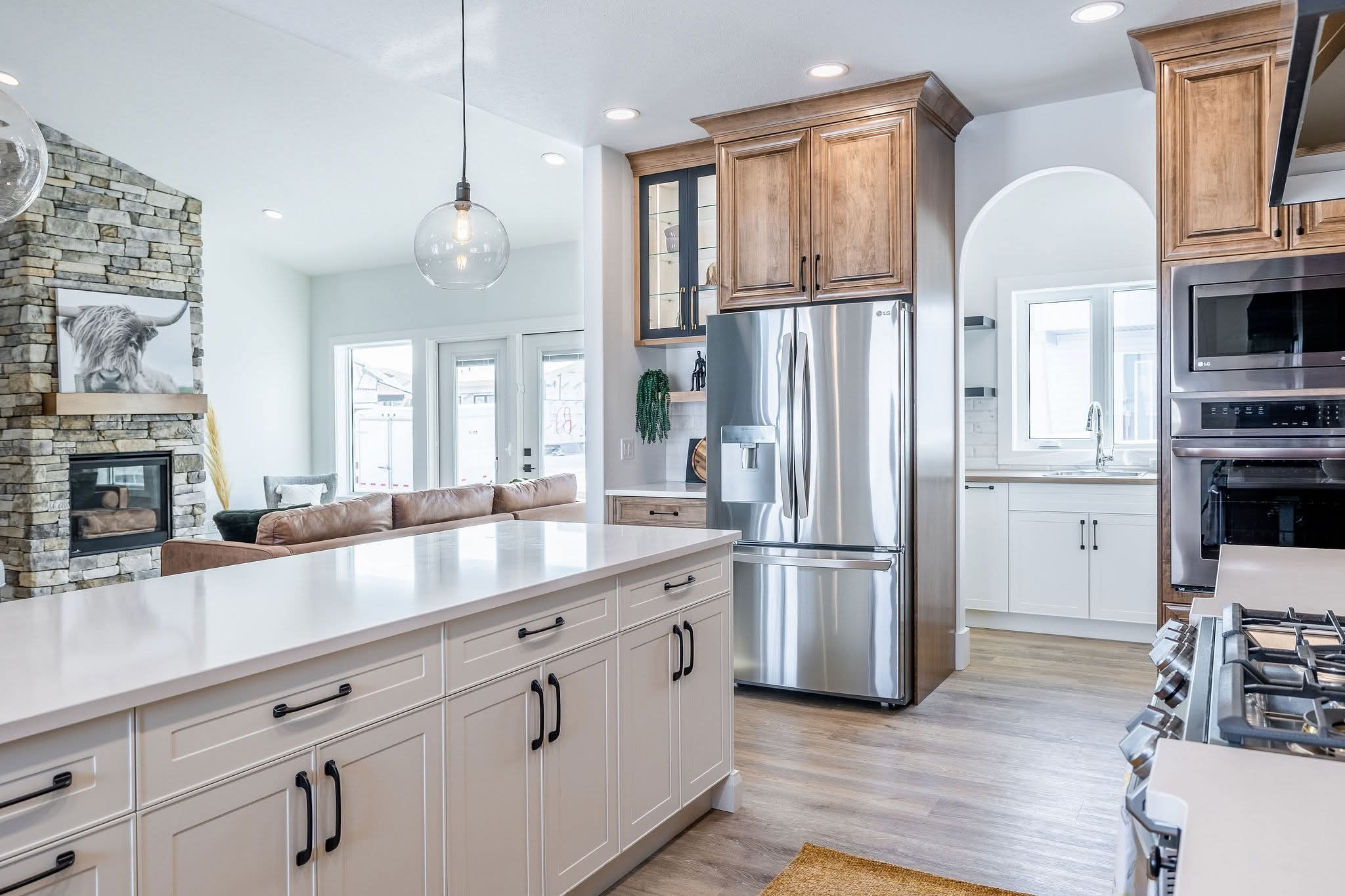 Modern kitchen with white cabinets, stainless steel appliances, and wooden accents. A cozy living room with a stone fireplace is visible in the background.