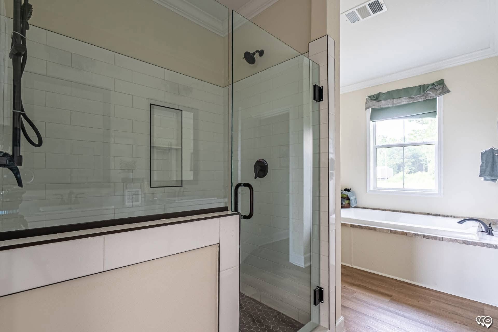 A modern bathroom with a glass-enclosed shower featuring white tiles and a dark handle. Nearby is a sunlit bathtub beneath a window with a green valance.