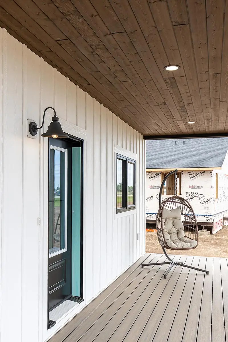 Covered porch with white siding and wooden ceiling. Features a hanging egg chair and black door. Construction visible in the background. Peaceful ambiance.