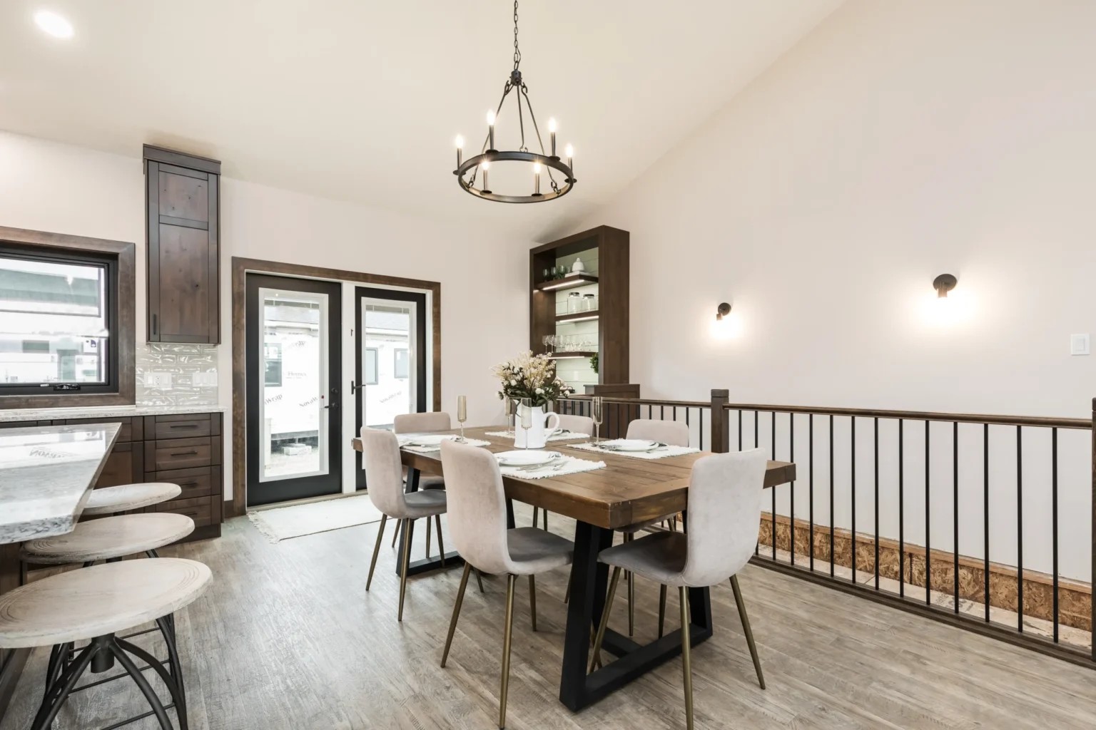 Bright dining room with a wooden table set for four, surrounded by beige chairs. A modern chandelier hangs overhead, with large windows and minimalist decor.
