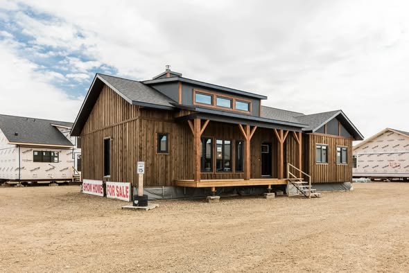 Wooden house for sale with large windows, small porch, and a rustic design under a cloudy sky. A "show home for sale" sign is visible.