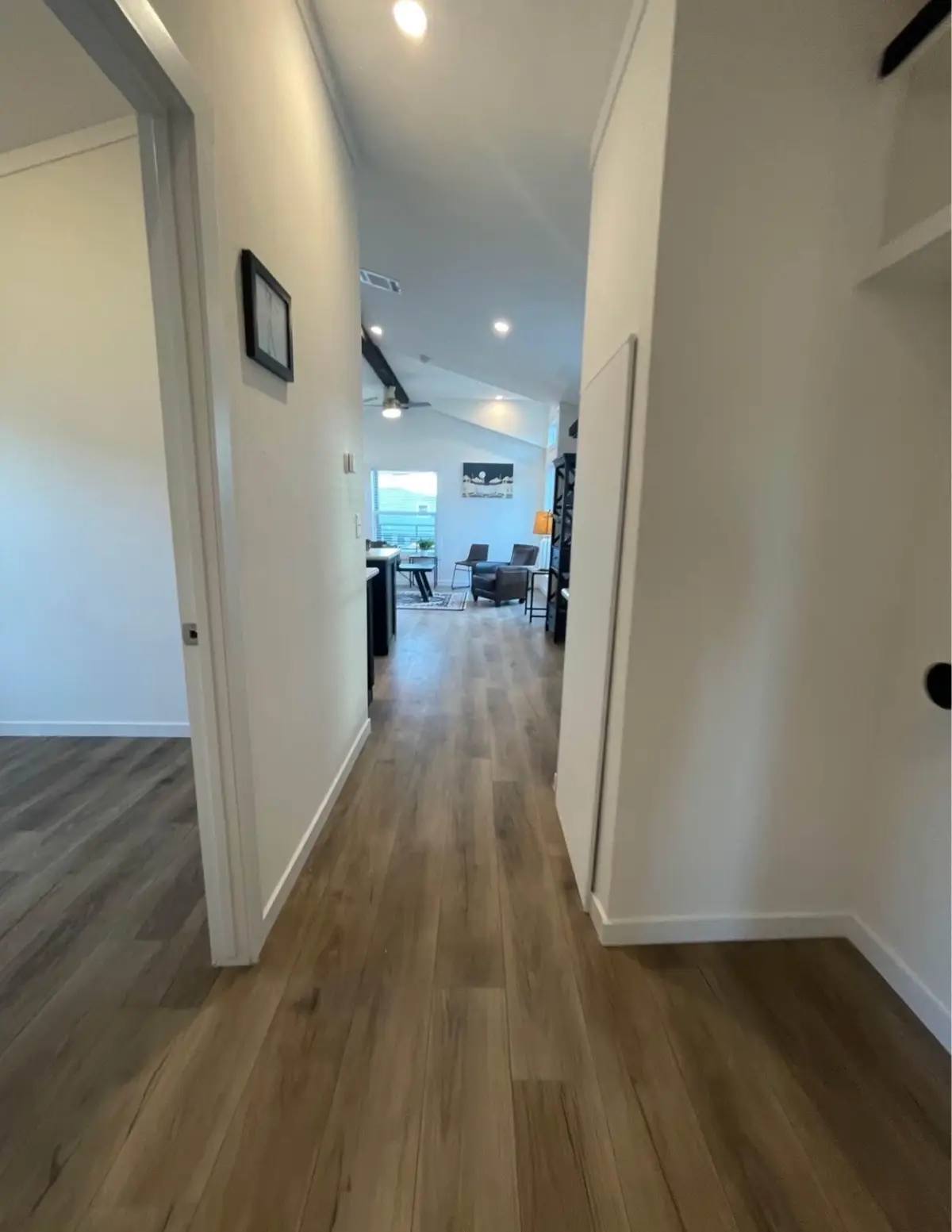 A modern hallway with wooden flooring leads to a bright living area. The space is minimalistic with a few chairs, a wall art piece, and natural light.