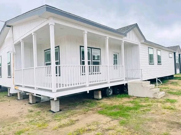 A modern white manufactured home with a small porch and railings. It sits on a grassy plot with concrete steps leading to the entrance, under a cloudy sky.