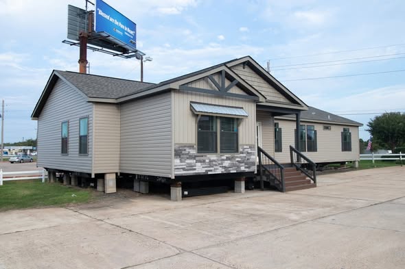 Single-story manufactured home on concrete stilts, featuring a modern design with beige siding and a brick accent. Located near a billboard.