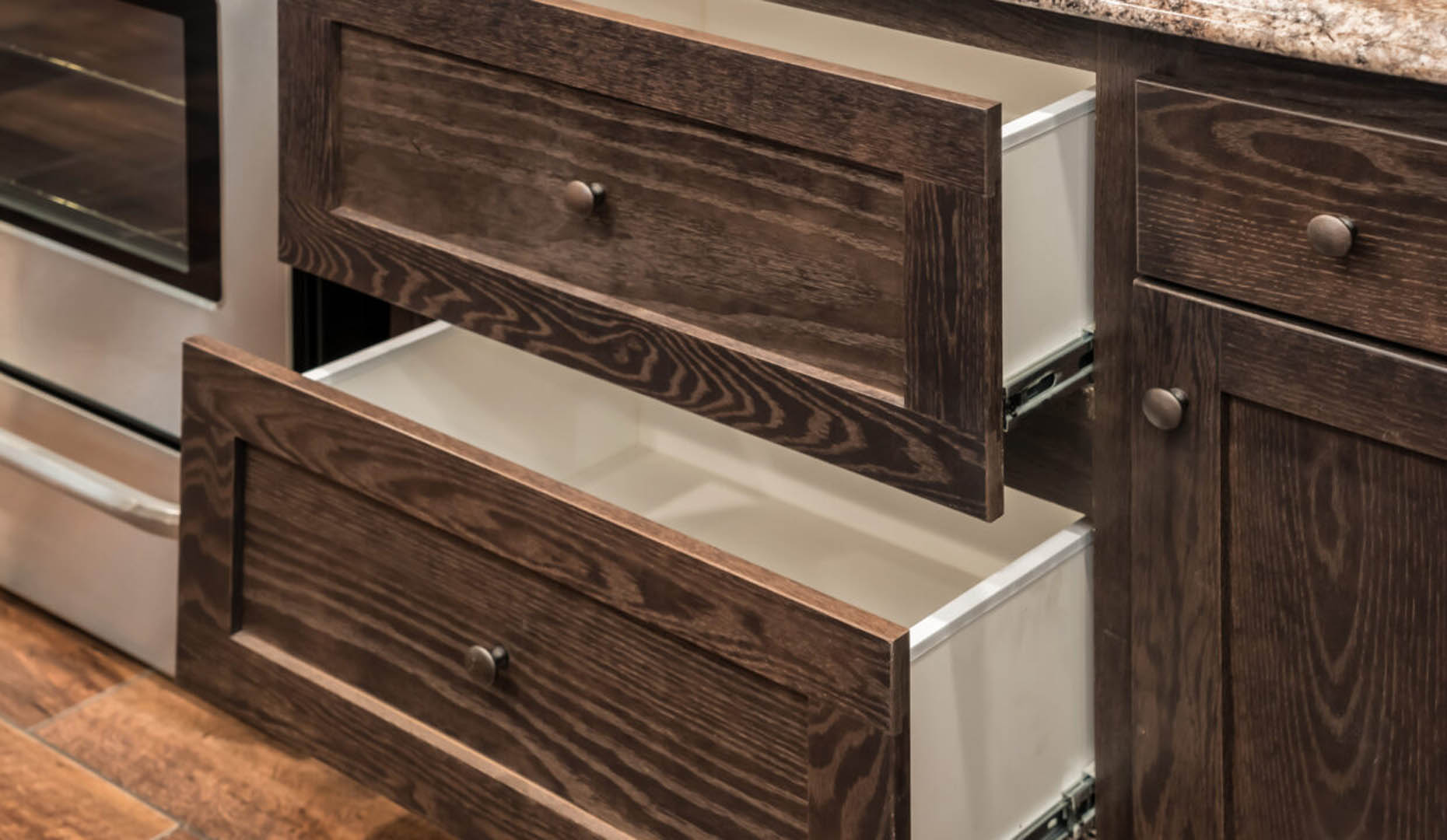 Open wooden kitchen drawers with a rich grain texture and dark knobs, alongside a stainless steel appliance and stone countertop, creating a warm, inviting tone.