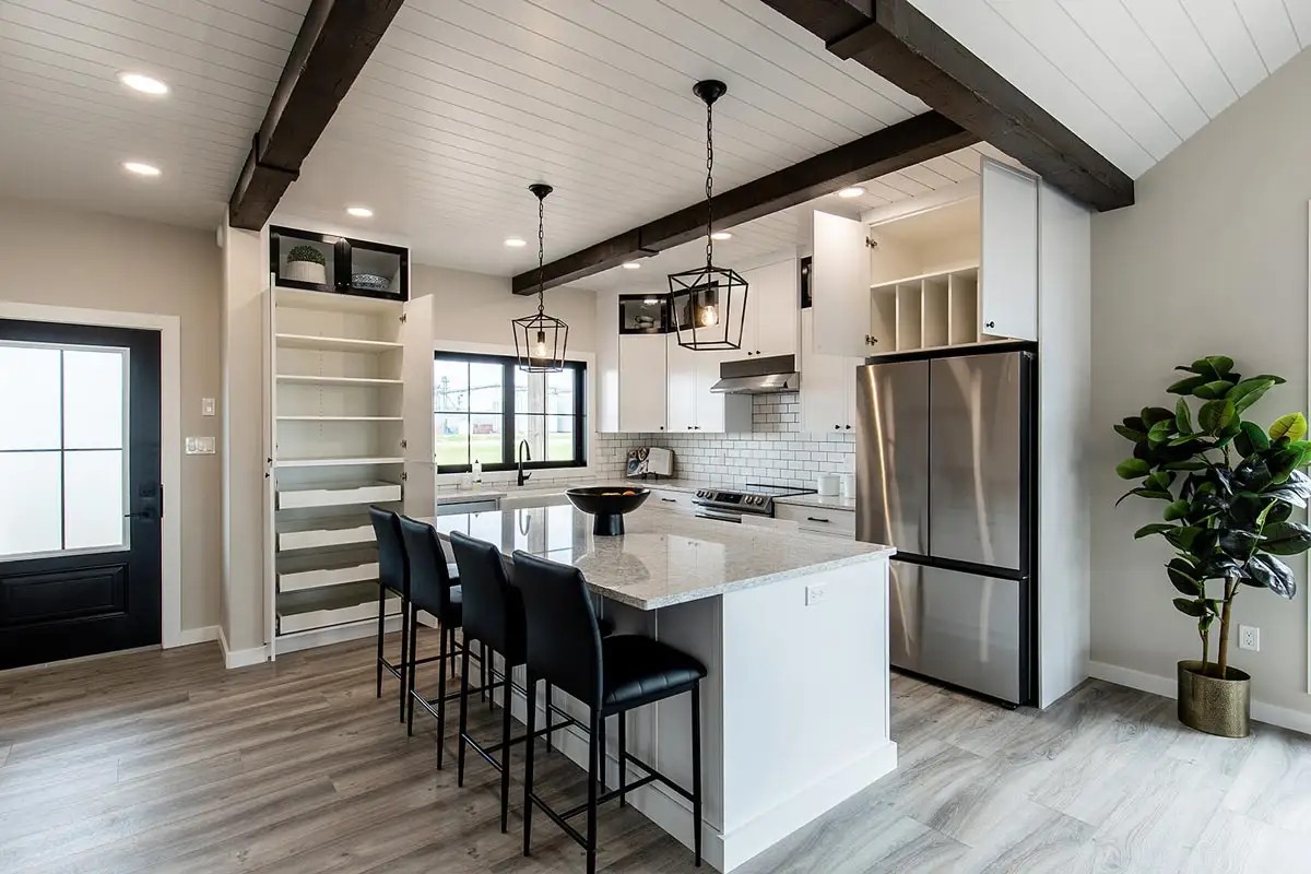 Modern kitchen with high ceiling and exposed beams, featuring a white island with granite countertop, black chairs, stainless steel appliances, and a potted plant.