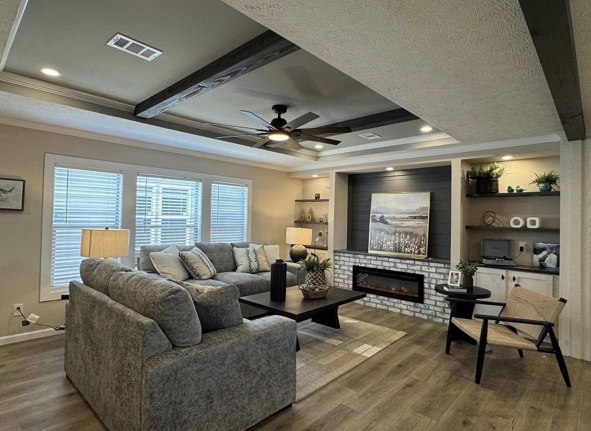 Modern living room with gray sectional sofa, large windows, and a ceiling fan. Brick fireplace with art, shelves, and warm lighting create a cozy ambiance.