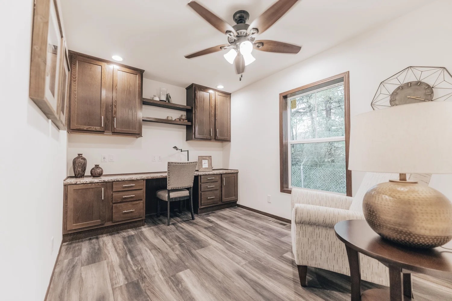 A cozy home office with dark wood cabinets, a built-in desk, and a cushioned chair. It features a window with a scenic view, a ceiling fan, and a lamp on a side table, creating a warm and inviting atmosphere.