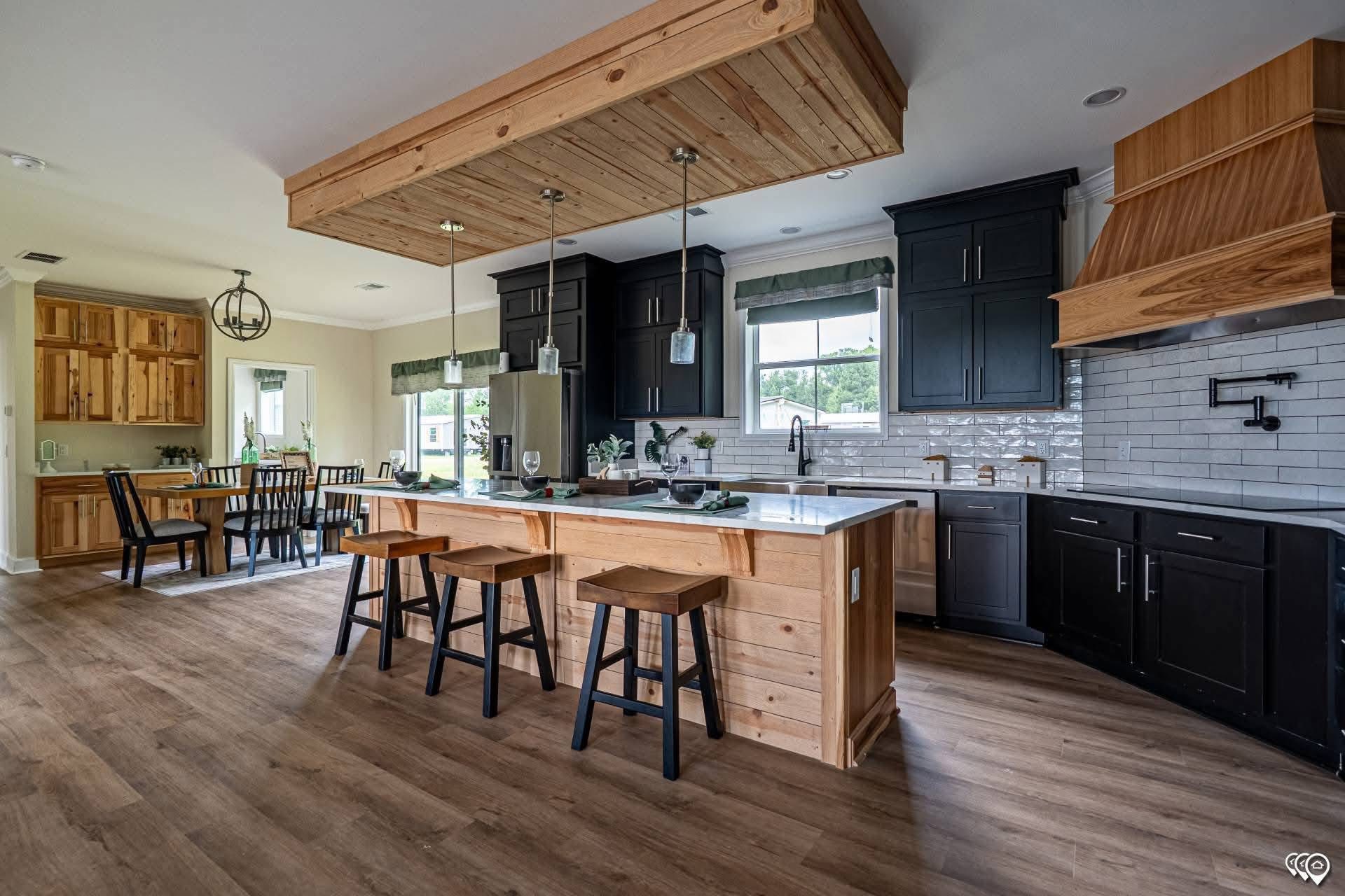 Spacious kitchen with a wooden island and stools, black cabinets, and a wooden ceiling feature. A dining area is visible with large windows, creating a cozy atmosphere.