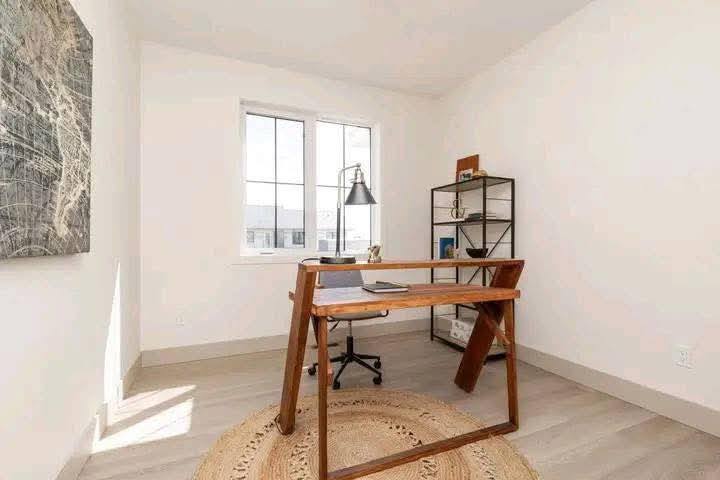 Minimalist home office with a wooden desk on a round rug, chair, and a lamp. Bright window and shelving unit with decor create a calm, airy ambiance.
