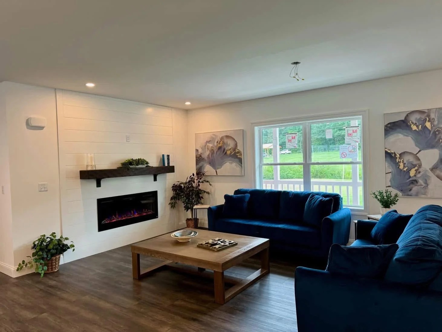 Modern living room with blue sofas, a wooden coffee table, and a sleek electric fireplace. Large window with lush outdoor view, abstract art on walls. Cozy and elegant.