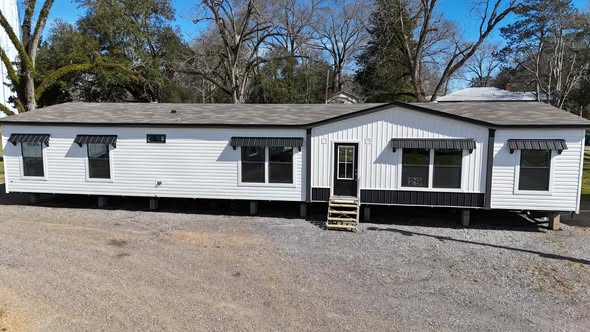 A large white manufactured home with black trim, six windows with awnings, and a small front porch. The home sits on a pebble lot surrounded by trees.