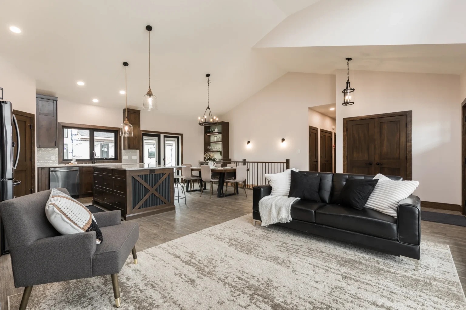 Open-concept living room and kitchen with neutral tones. Features a black sofa with white pillows, a gray armchair, a large rug, and modern lighting. Cozy and inviting.