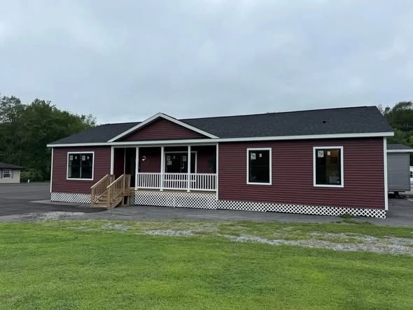 A single-story red building with white trim and a small porch. It has multiple windows and is set against a backdrop of green grass and cloudy sky.