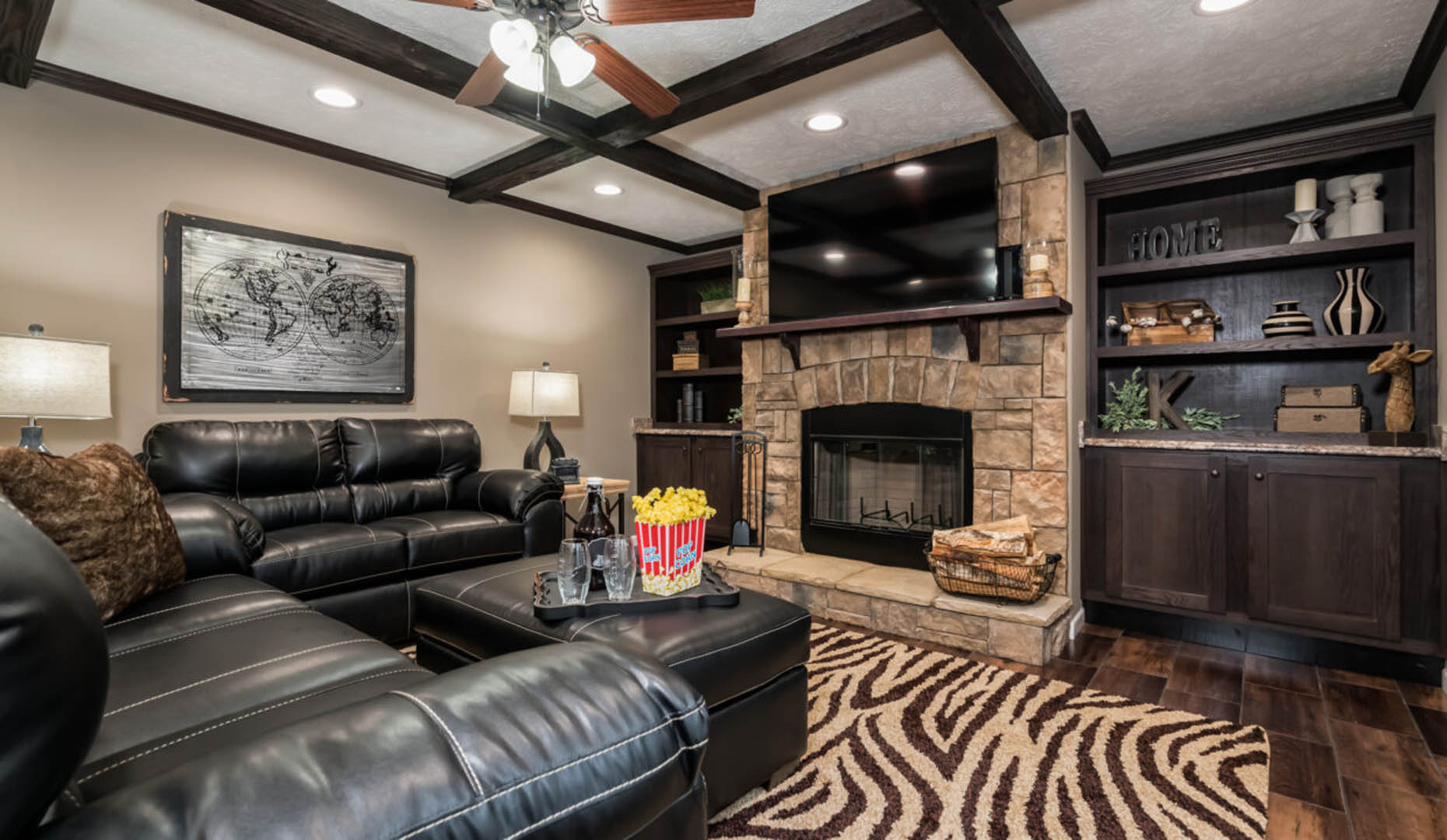 Cozy living room with black leather sofa, patterned rug, stone fireplace, and built-in shelves. Warm lighting and modern decorative elements create a welcoming atmosphere.