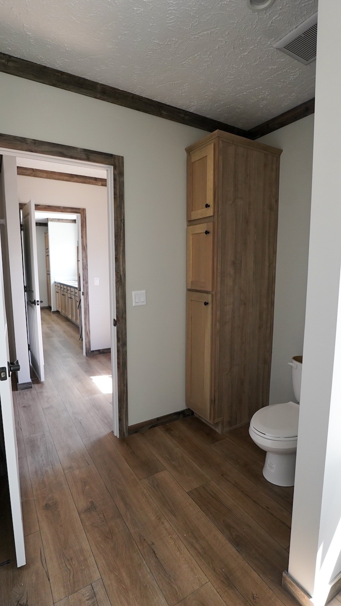 A small, minimalist bathroom with wooden floors and a tall wooden cabinet beside a white toilet. An open door reveals a hallway with similar decor.