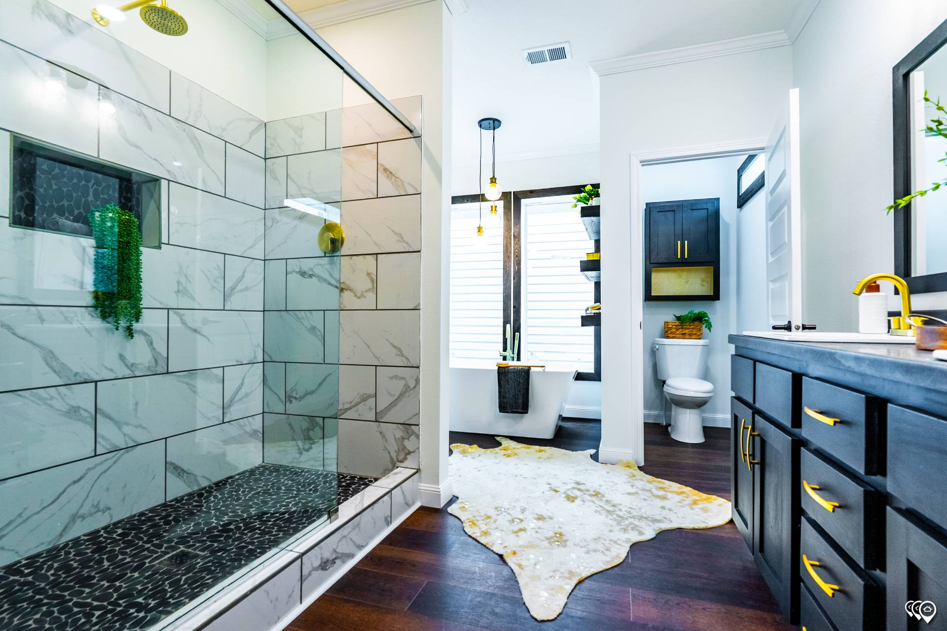 Modern bathroom with a marble-tiled shower, glass doors, dark wood floors, a freestanding tub, and a sleek vanity with gold accents. Bright and stylish.