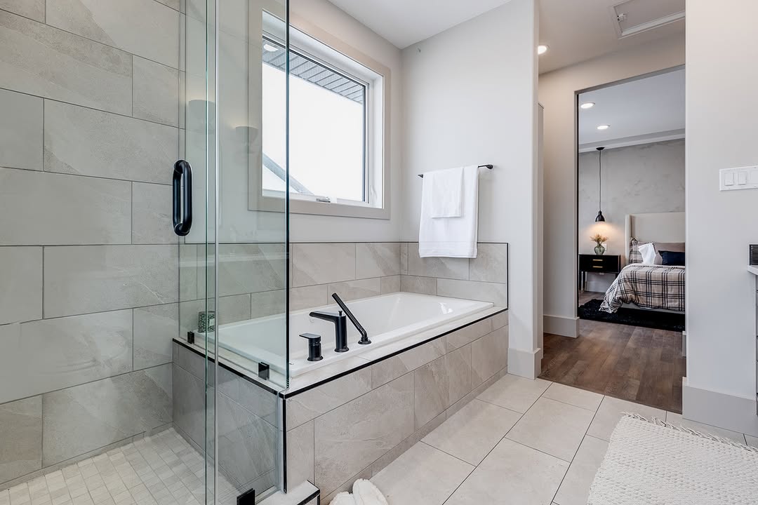 Modern bathroom with a glass shower, soaking tub, and black fixtures. A towel hangs nearby. The room opens to a cozy bedroom with wood flooring.