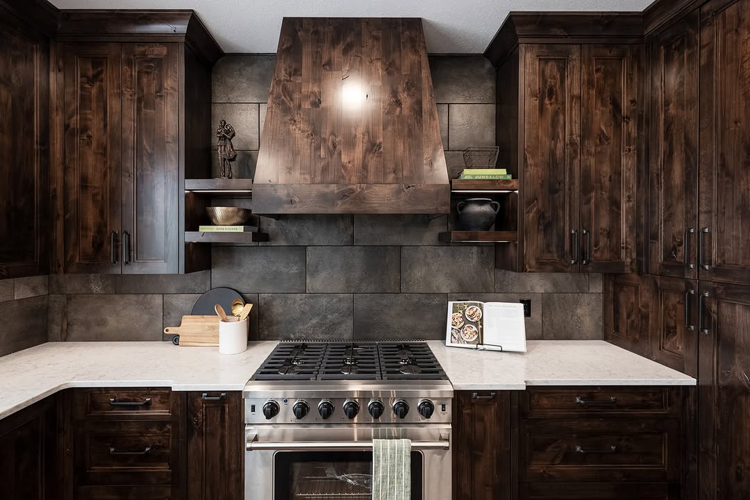 Warm, rustic kitchen with dark wood cabinetry, stainless steel oven, stone backsplash, and white countertops. Open shelves hold books and decor.