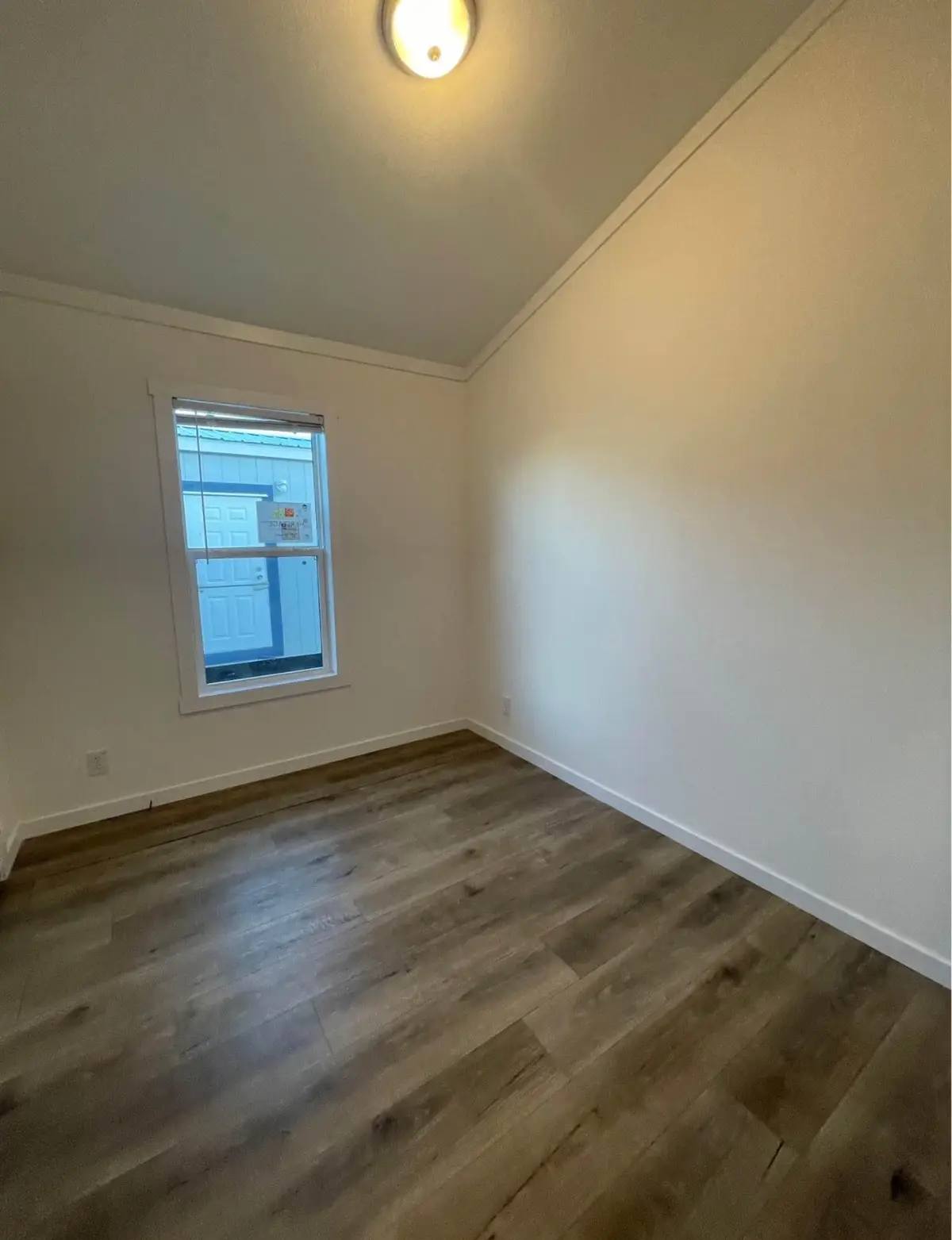 Small empty room with beige walls, a single window, and wood laminate flooring. Ceiling light fixture adds warmth. Calm, minimalist atmosphere.