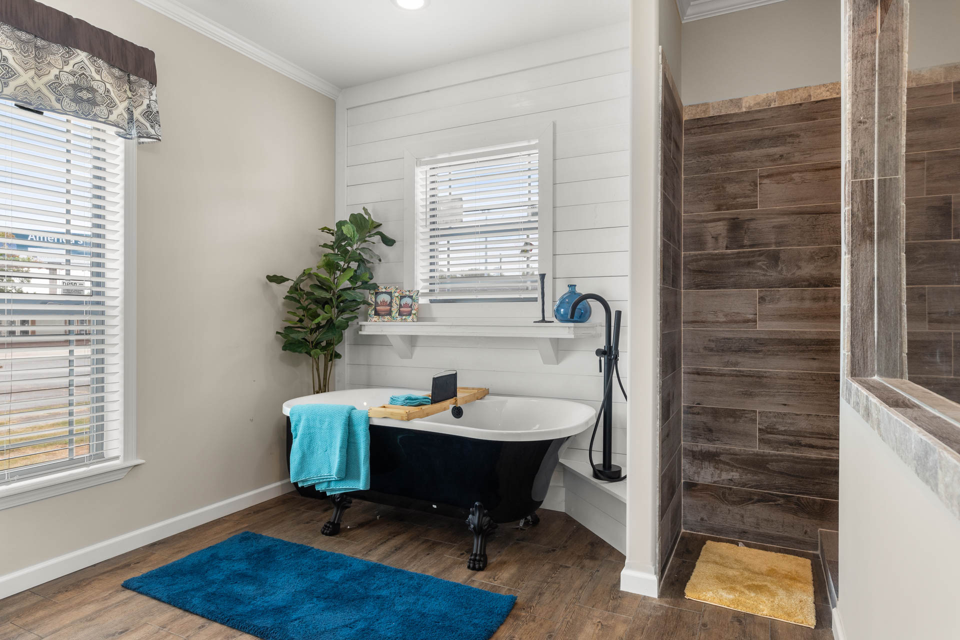 Stylish bathroom with a black clawfoot tub, wooden flooring, blue mat, and towel. Natural light from windows, plant, and rustic wood accents create a serene atmosphere.