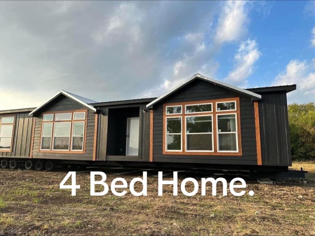 Modern modular home with dark siding and large windows, set in a grassy area under a cloudy blue sky. Text reads "4 Bed Home," suggesting spaciousness.