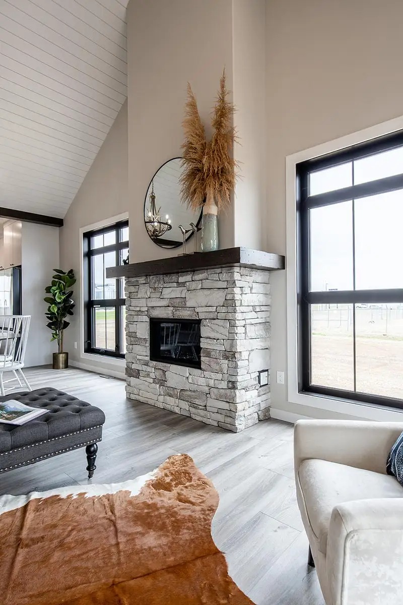 Modern living room with stone fireplace, large windows, and beige walls. Features a round mirror, pampas grass, and a cozy chair. Bright, airy ambiance.