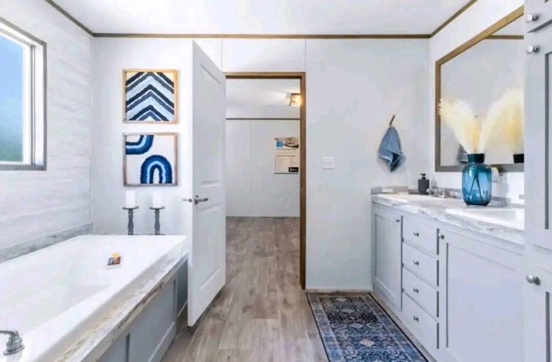 A modern bathroom with light gray walls and wood flooring. Features include a bathtub, white cabinets, blue and white abstract art, and a blue vase with pampas grass.