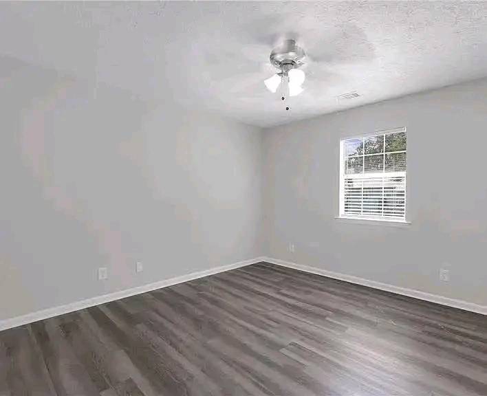A bright, empty room with light gray walls and sleek, dark hardwood floors. A ceiling fan with lights hangs overhead, and sunlight streams through a window with blinds, creating a serene, clean atmosphere.