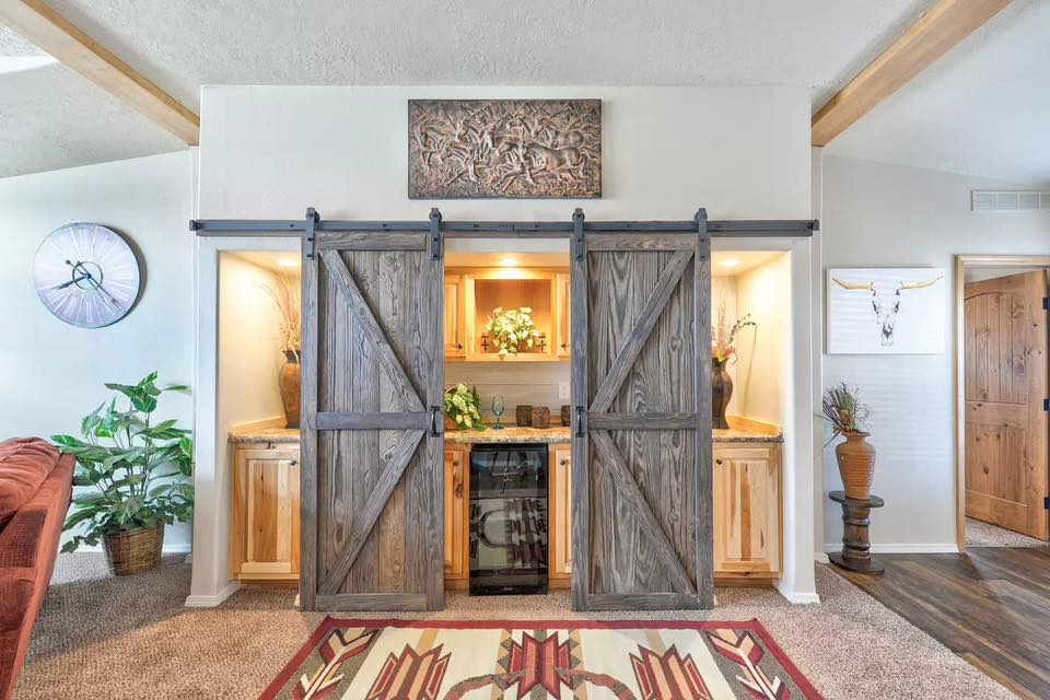 Open-concept ranch home interior with a rustic kitchen featuring oak cabinets, granite countertops, a farmhouse dining area with horse-themed decor, a spacious living room with vaulted ceilings and red sofas, and a master bath with double vanities and barn doors.