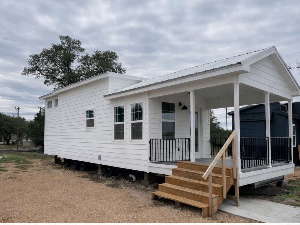 White tiny home with a front porch features three steps, black railings, and multiple windows. It sits on a gravel lot under a cloudy sky, surrounded by trees.