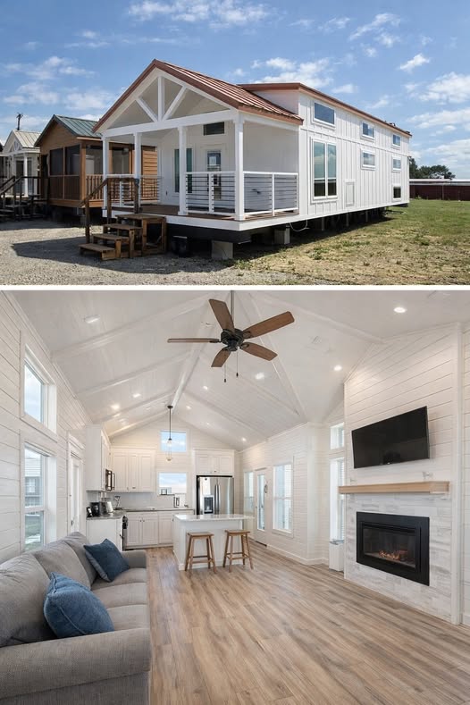 Alt text: "A modern tiny house with white siding and a red roof is set on a grassy lot, featuring a small porch. Inside, a bright, open-plan living area with vaulted ceilings, a ceiling fan, wood flooring, and white walls includes a cozy gray sofa, wall-mounted TV, and electric fireplace. The kitchen area has white cabinetry, stainless steel appliances, and a small island with barstools, creating a welcoming, minimalist atmosphere."