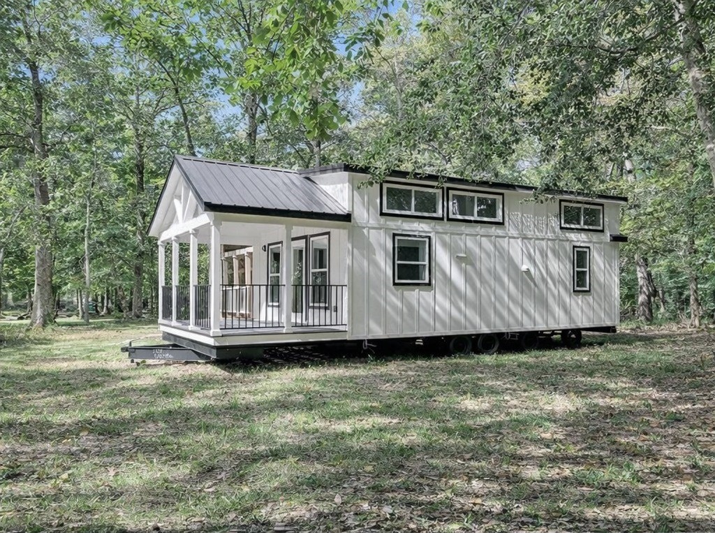 A modern tiny house on wheels sits in a lush, wooded area. It has a white exterior, large windows, and a small porch. The scene feels peaceful and natural.
