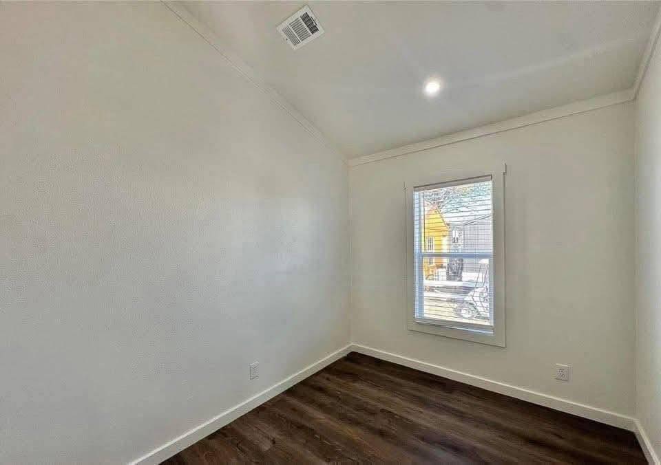 Small, empty room with white walls, dark wood flooring, and a single window. Natural light illuminates the space, creating a bright and minimalistic ambiance.