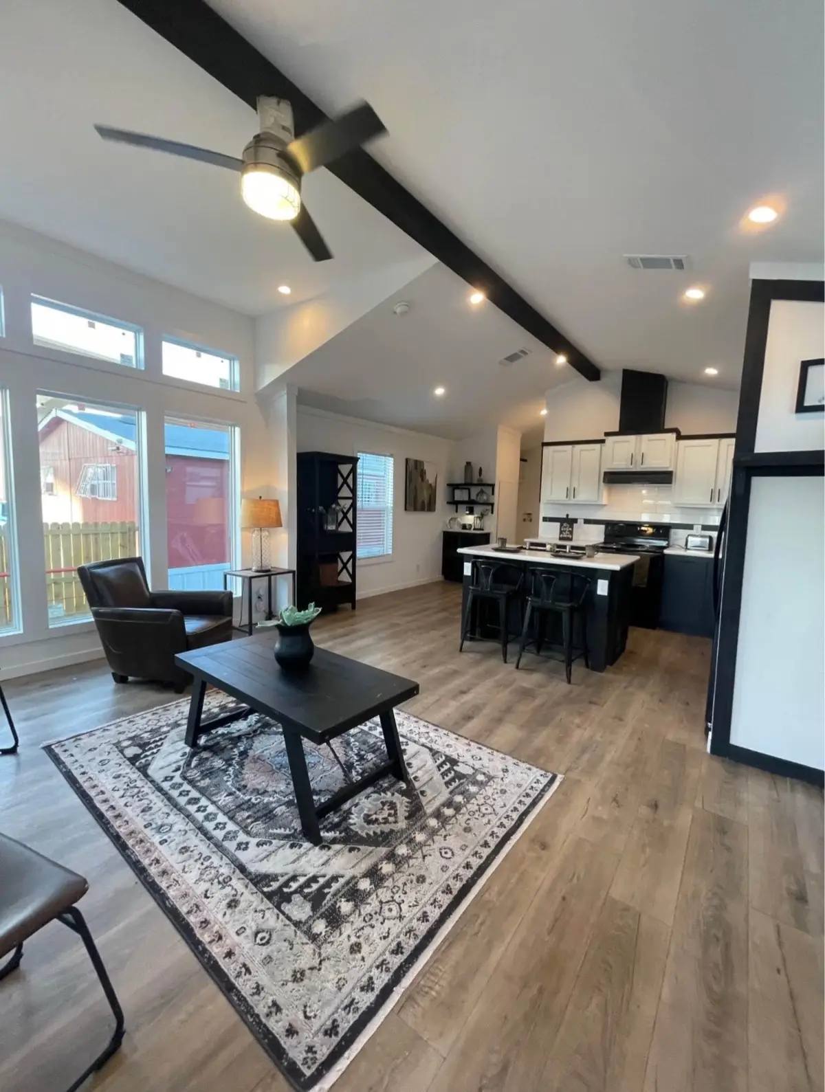 Spacious living room with high ceilings and wooden floors. Features a patterned rug, modern coffee table, leather armchair, ceiling fan, and open kitchen.