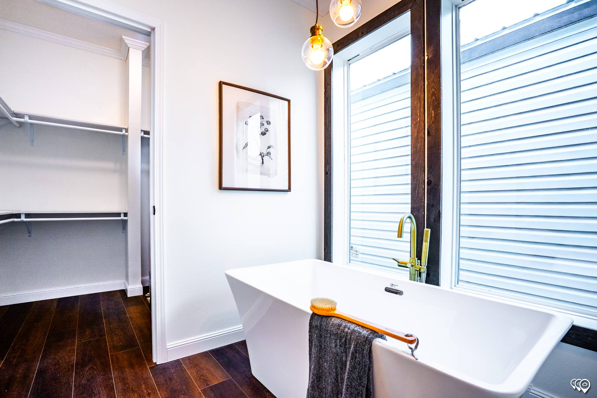 Modern bathroom with a white freestanding bathtub, brass faucet, and towel. Large windows with natural light and a minimalist framed photo.