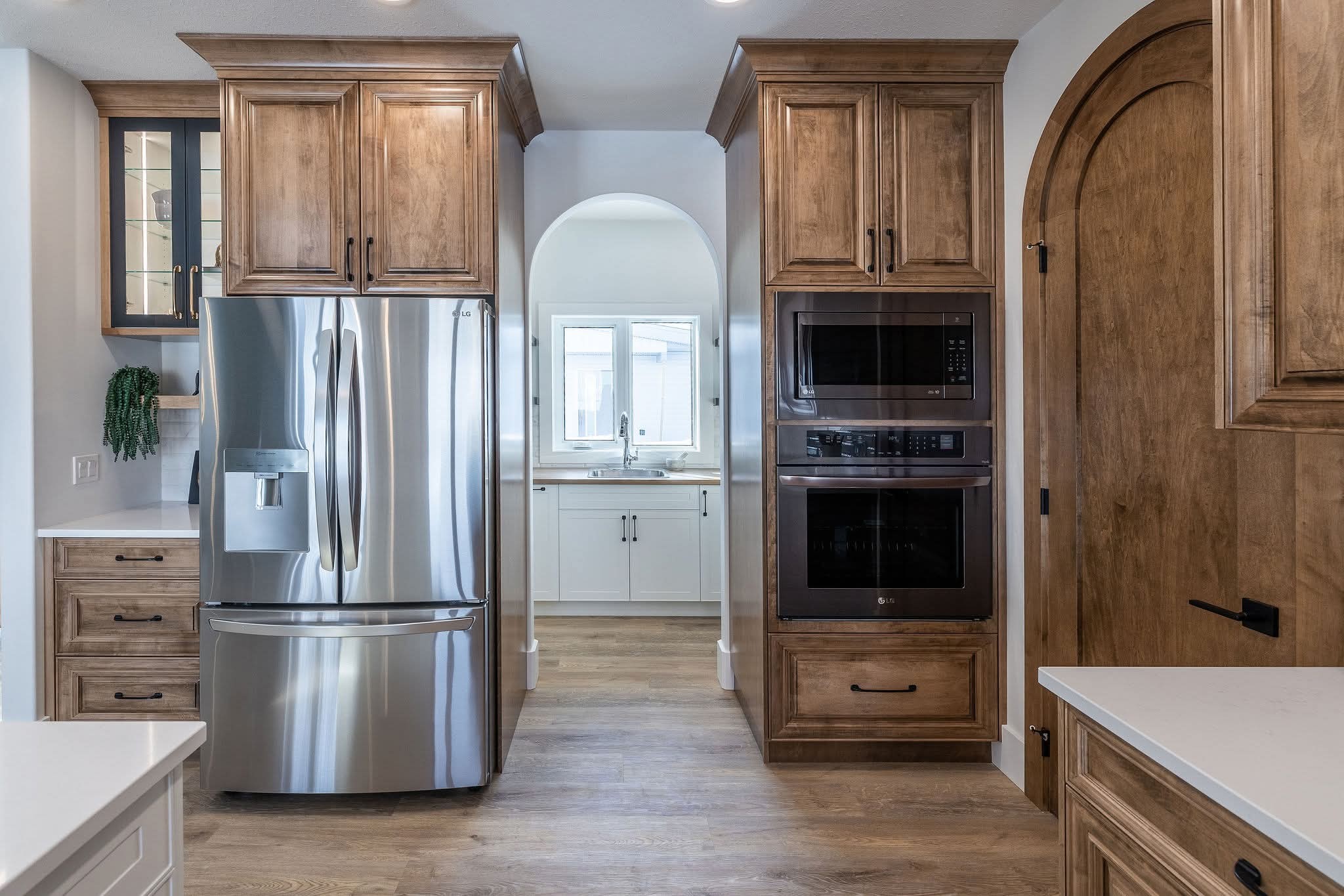 Modern kitchen with stainless steel fridge, wooden cabinets, and a double oven. An archway reveals a bright sink area with a window. Warm and inviting.