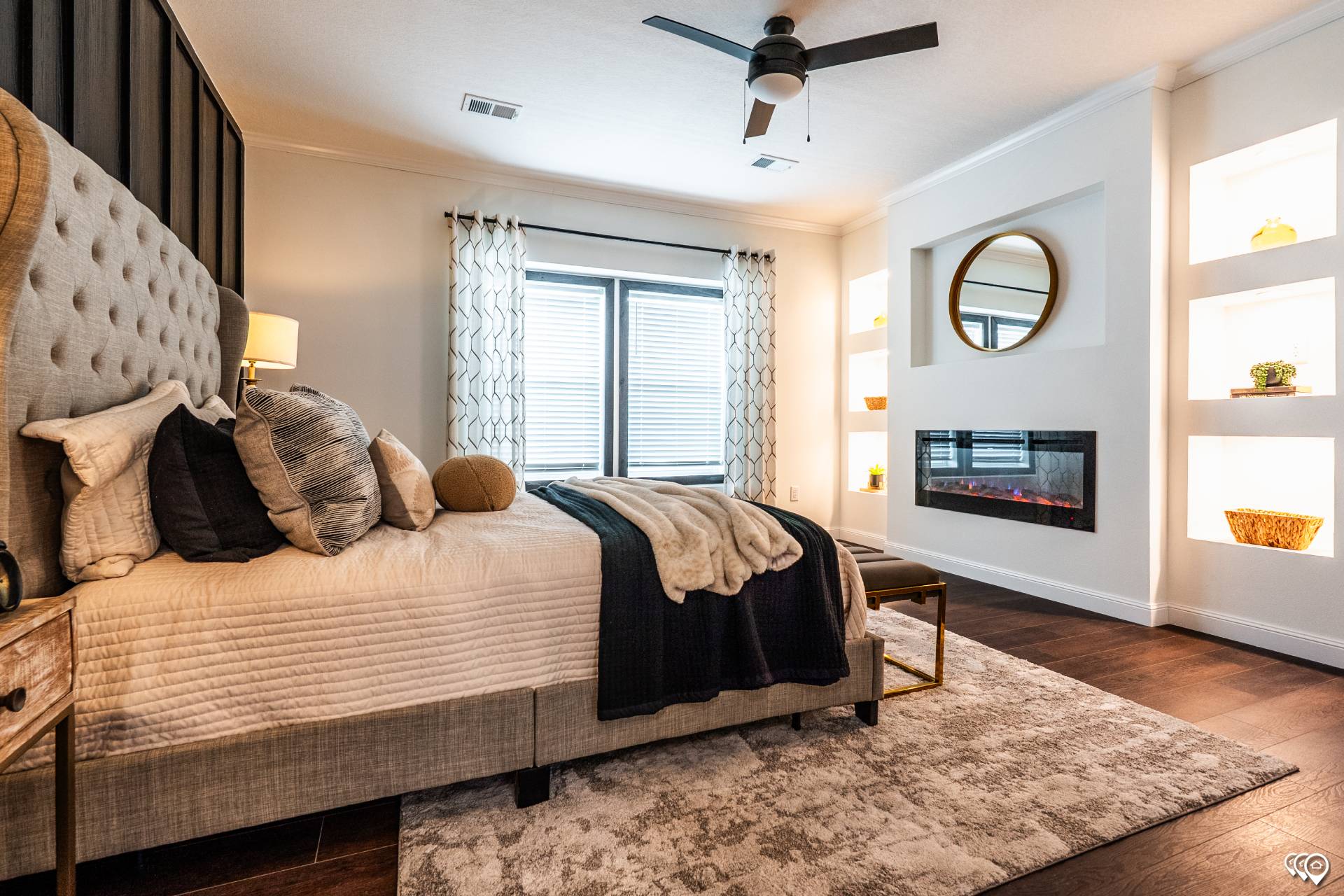 Modern bedroom with a plush beige bed and pillows, next to a side table with a lamp. A large window with patterned curtains lets in soft light. A wall-mounted electric fireplace sits beneath a round mirror, flanked by lit shelves with decor. The room has warm tones and a cozy, elegant atmosphere.