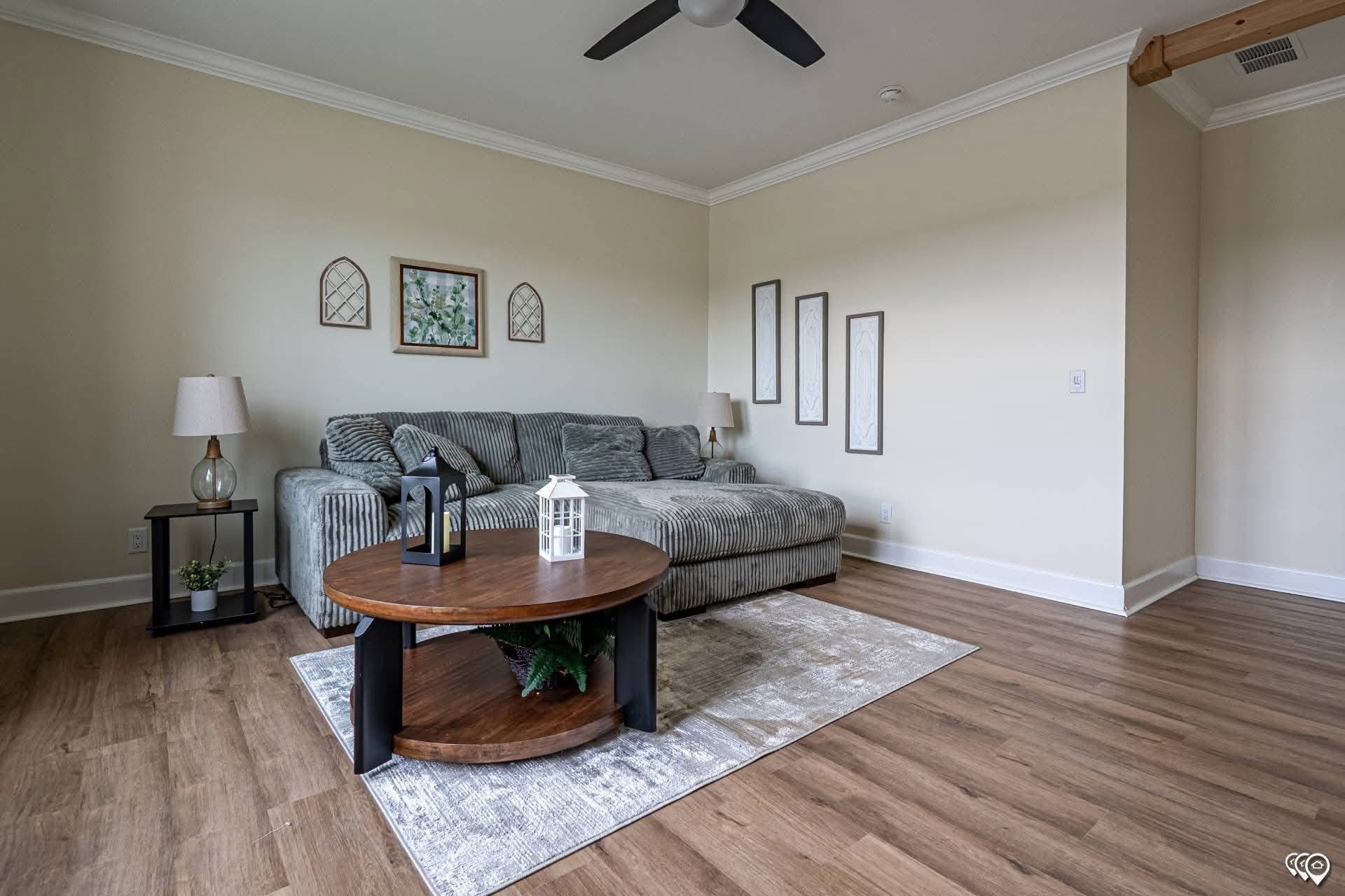A cozy living room featuring a striped gray sectional sofa with decorative pillows, a round wooden coffee table, and wall art. The room has a warm and inviting feel with wood flooring and a ceiling fan above.
