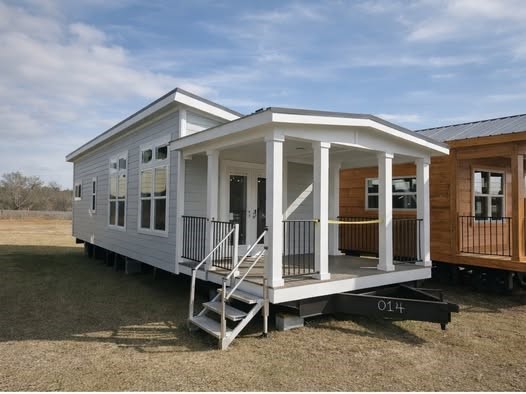 A small, white prefabricated home with a covered porch and steps sits on grassy land under a partly cloudy sky. Emphasizes a clean and simple design.