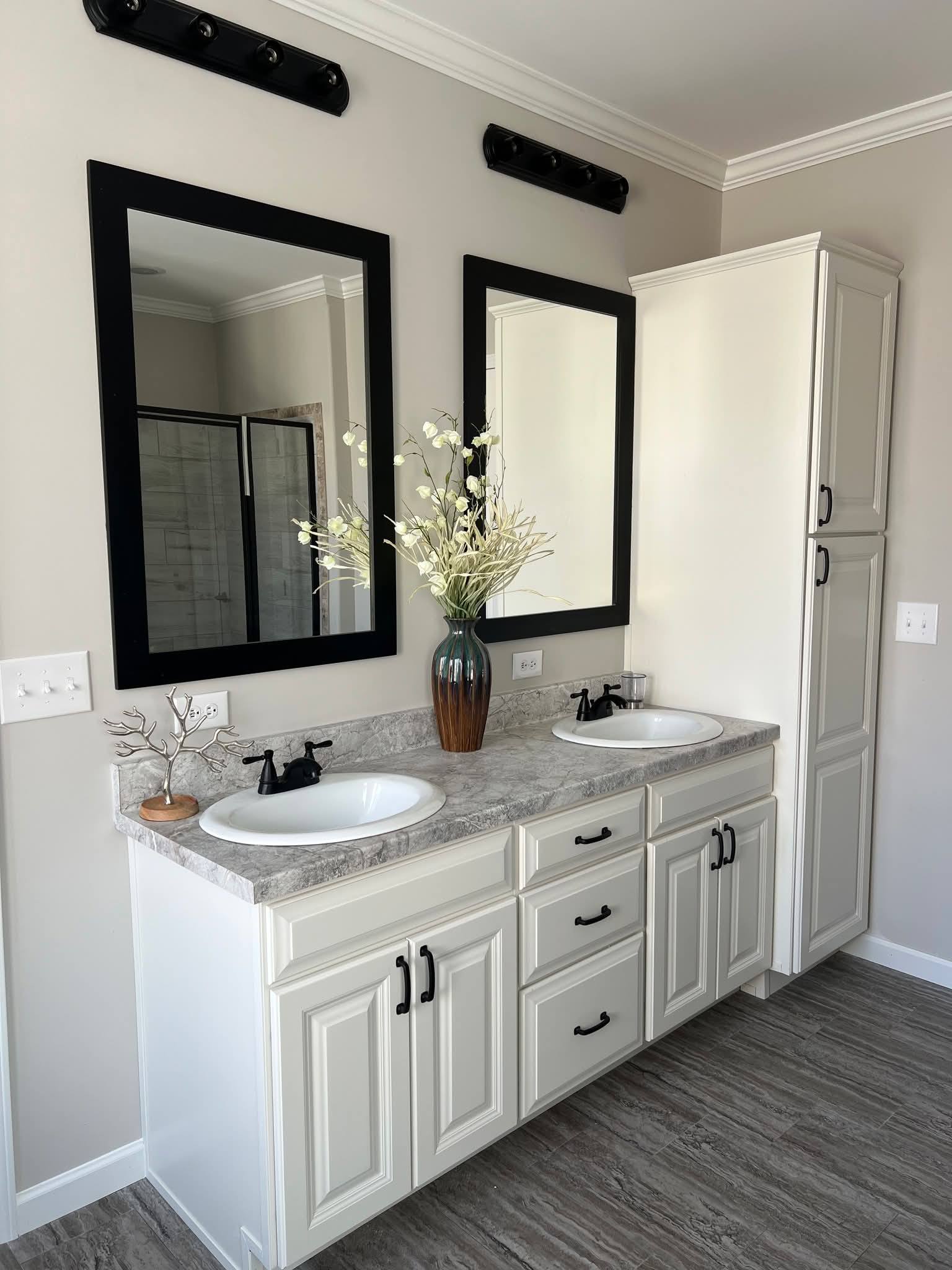 Modern bathroom with dual sinks and granite countertop. Two rectangular mirrors with black frames hang above, and a vase with white flowers adds elegance.