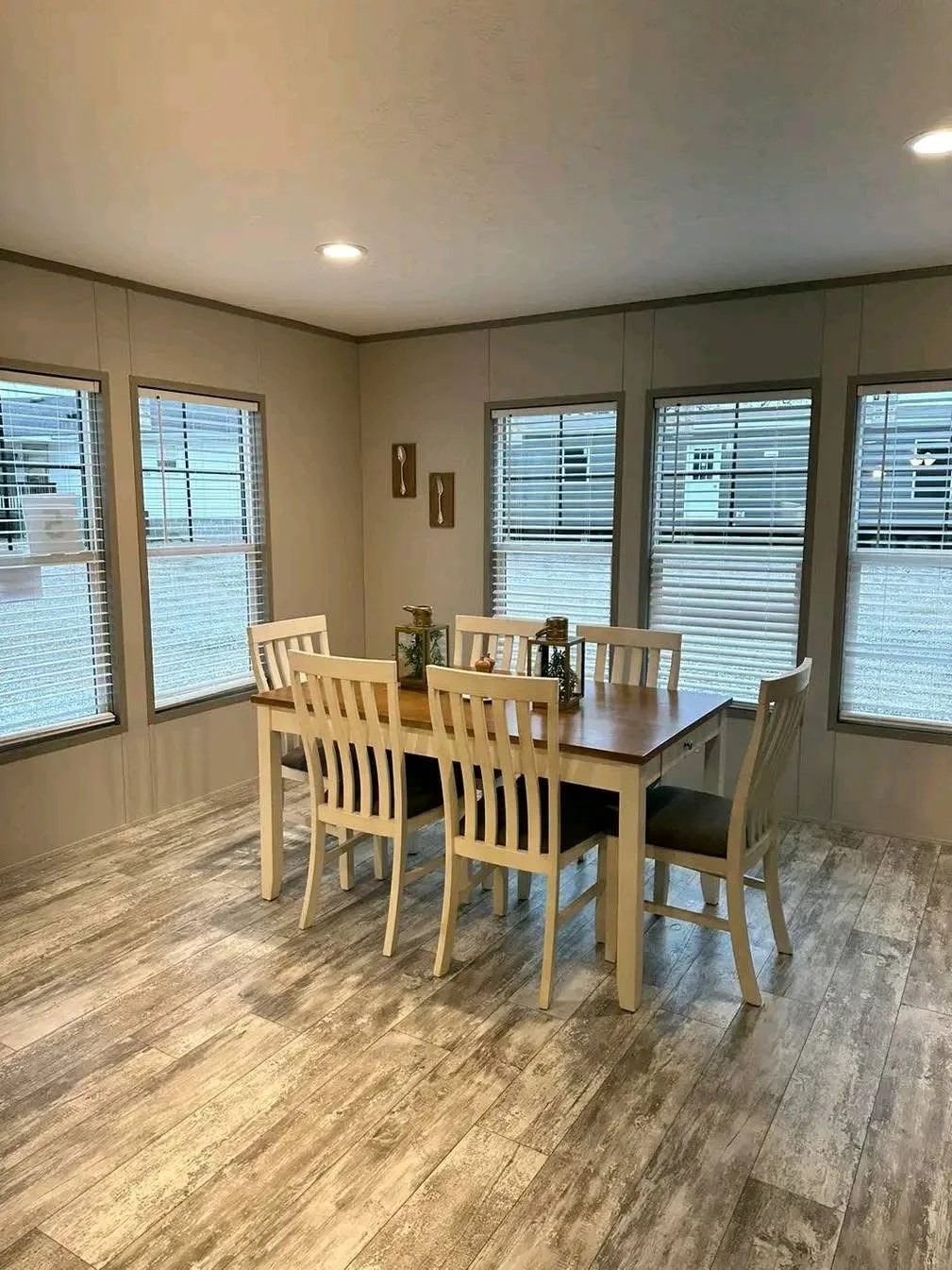 A bright dining area featuring a rectangular table with a light wood top and white legs, surrounded by six matching white wooden chairs with grey seats. The room has grey wood-look flooring, neutral-toned walls, and multiple large windows with white horizontal blinds."