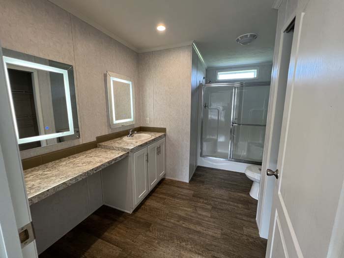Luxury manufactured home master bathroom featuring a double vanity with LED-lit mirrors, marble-style countertops, and a glass-door walk-in shower.