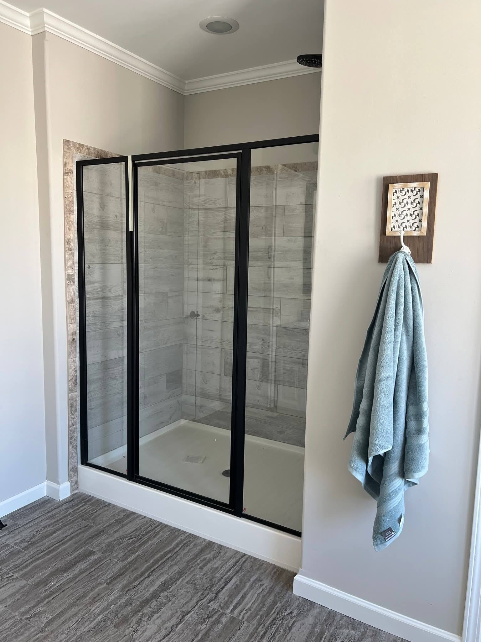 Modern bathroom with a glass shower enclosure featuring black frames and light gray tiles. A blue towel hangs on a wall hook, giving a serene feel.