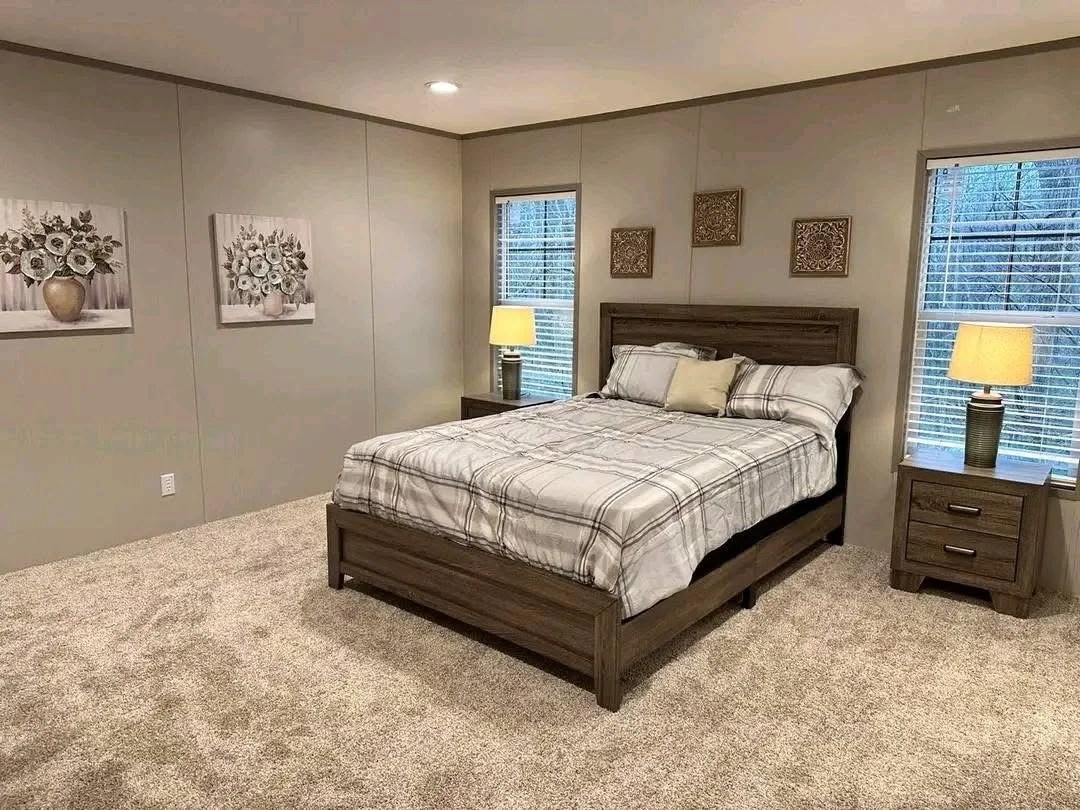 "A modern bedroom featuring a dark wood bed frame with a grey and white plaid comforter. The room has plush tan carpeting, neutral grey walls decorated with floral artwork, and two matching nightstands with warm bedside lamps beside windows with white blinds."