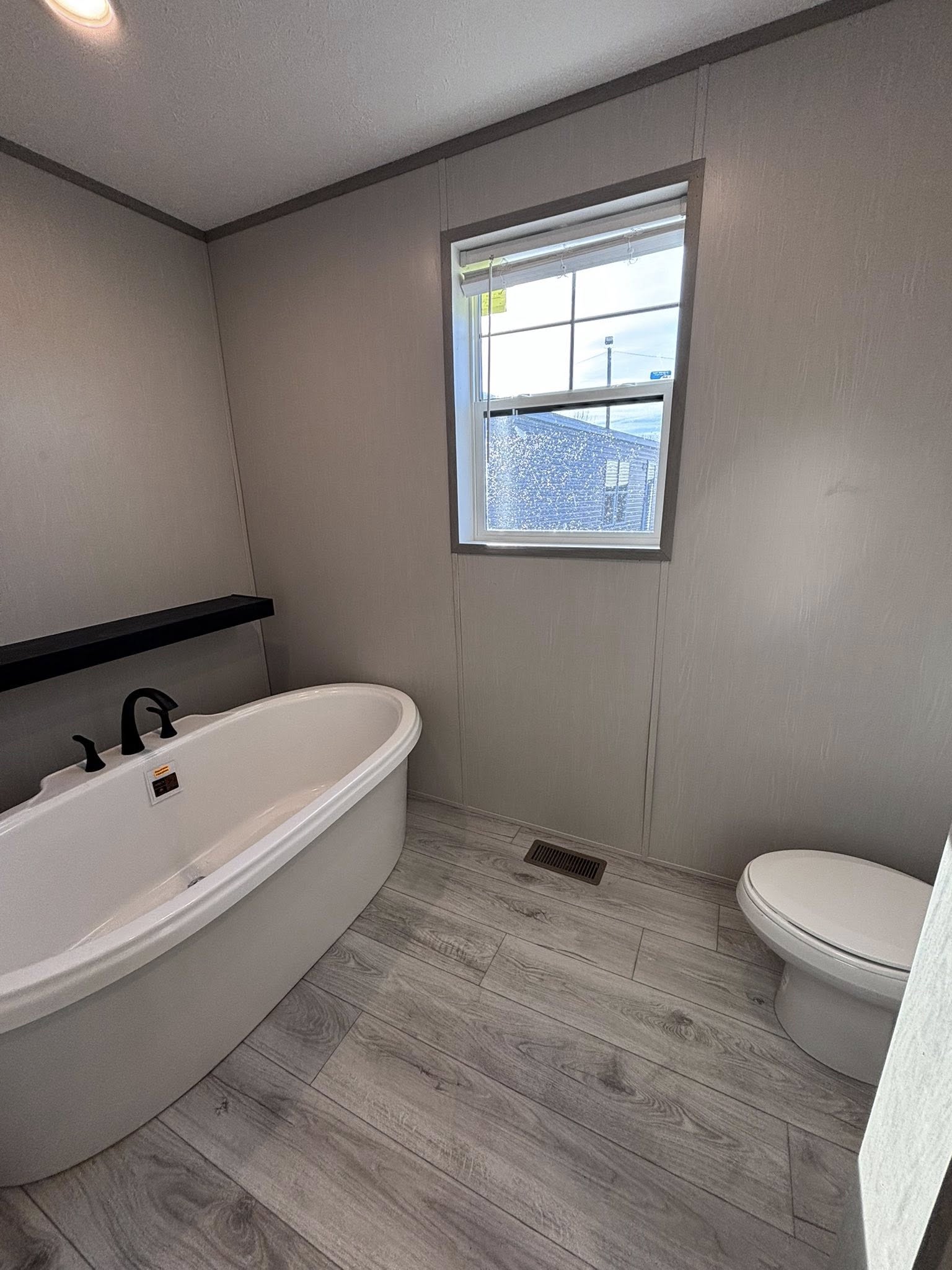 A collection of images showcasing a modern farmhouse-style interior with light wood-plank flooring and a neutral color palette. The dining area features a dark wood-top table with white legs, white chairs, a matching bench, and rustic wall decor including a large forest path canvas and a decorative wooden fork. The kitchen is equipped with grey cabinetry, a large island with a swirling marbled countertop, cushioned barstools, and an industrial-style chandelier with Edison bulbs. Functional details include a white farmhouse sink, stainless steel appliances, and a sliding wooden barn door. The bathroom sections highlight a white freestanding tub, a double vanity with a marbled top, and dark cabinetry against light grey walls.