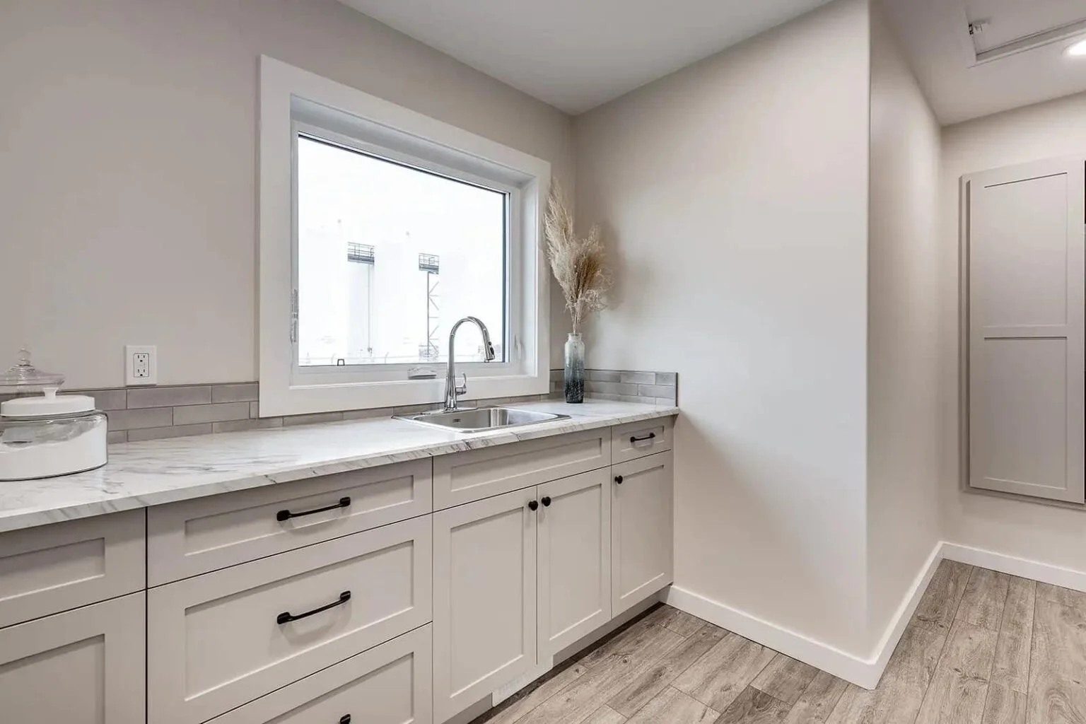 A modern laundry room features light beige cabinets, a marble countertop, and a large window. A vase with dried plants adds a touch of elegance. Warm and tidy.